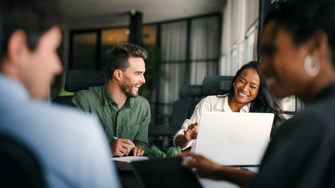 Colleagues laughing together around a meeting table with an open laptop